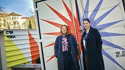Zwei Frauen vor einem Container, der mit grafischen Elementen gestaltet ist.