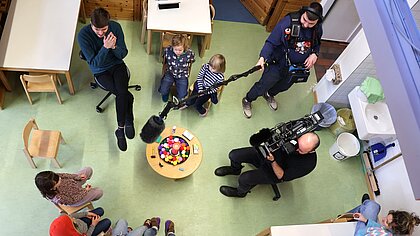 Ein Erzieher und Kinder sitzen in der katholischen Kita Wilde Wanne im Kreis, ein SWR-Team filmt