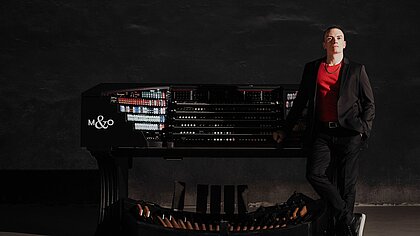 Cameron Carpenter neben seinem Instrument. Dunkles Bild, nur Orgeltasten und rotes T-Shirt leuchten